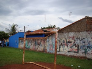 Foto mostra gol do campo de futebol e ao fundo a casa da moradora que é atingida pelas bolas